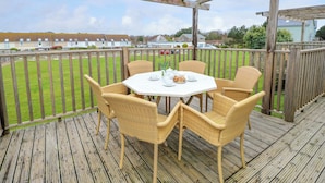 A wooden deck with wicker chairs and a table set for a meal, overlooking a grassy area and houses. - Munday H2 (Yarmouth)
