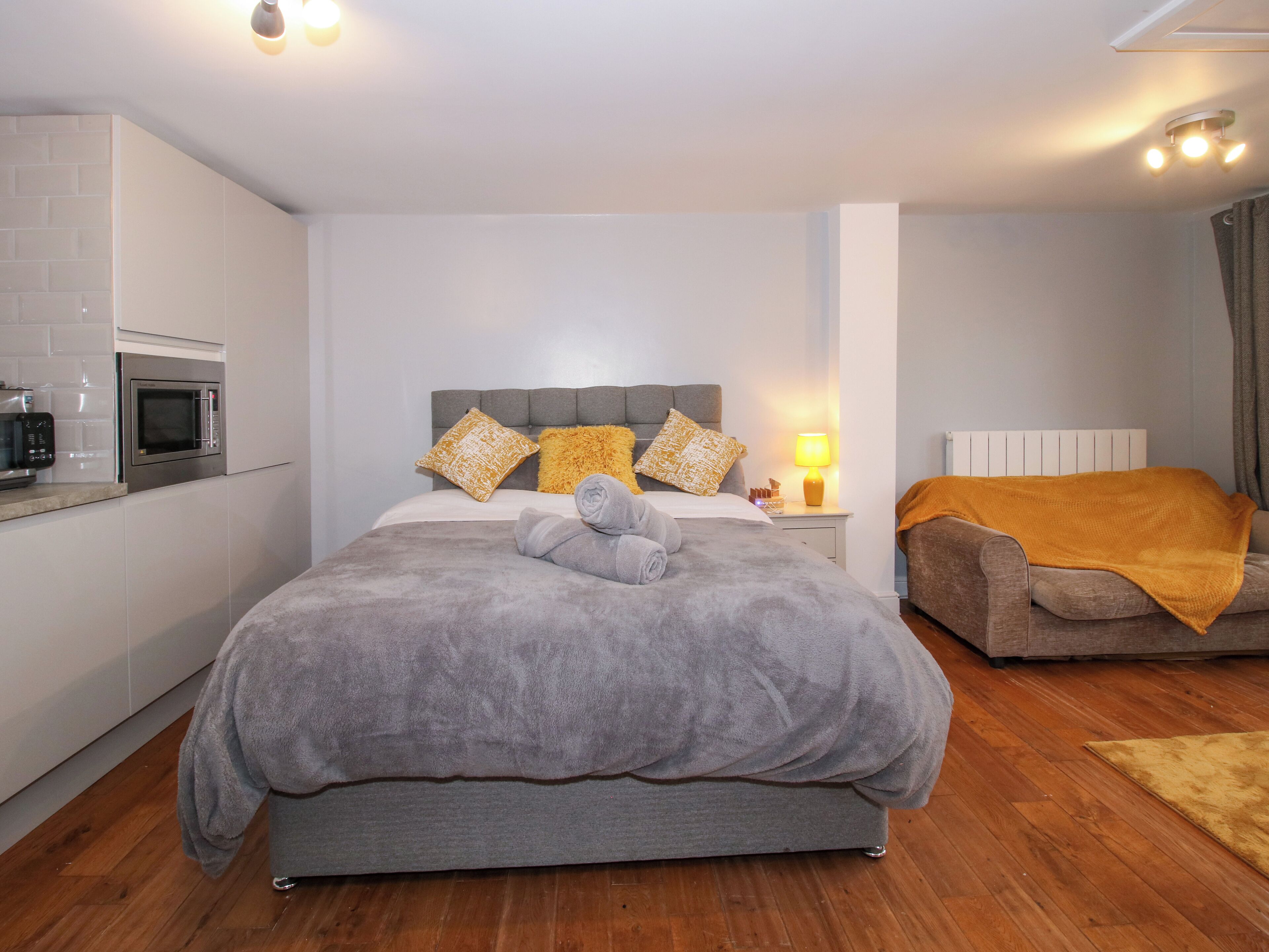 A bedroom with a bed, a bedside table, a lamp, a sofa, and a microwave in the kitchen area.