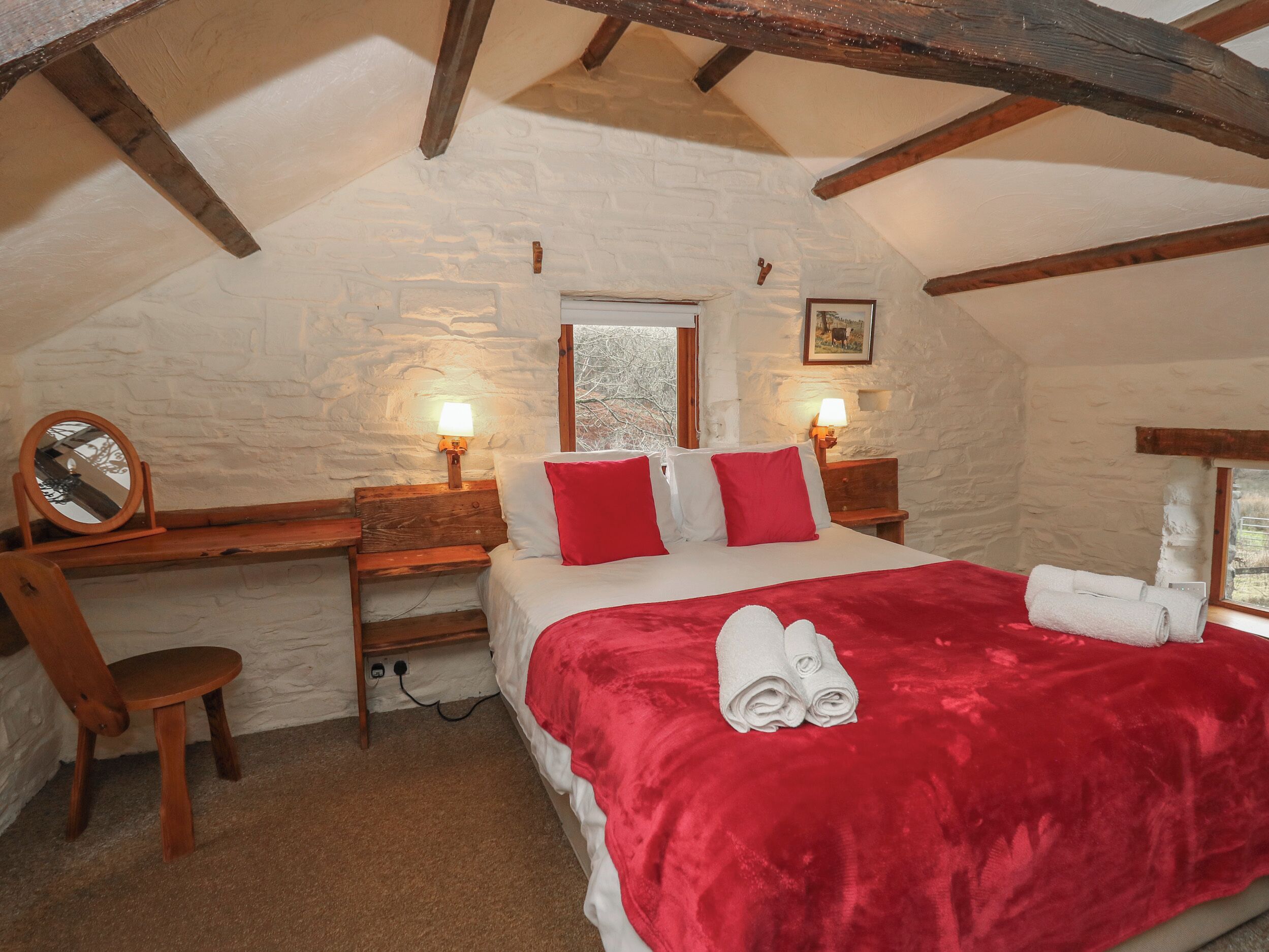 A cozy bedroom with a stone wall, a wooden bed with red bedding, a bedside table with a lamp, and a chair.