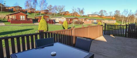 Eine Holzterrasse mit Tisch und Stühlen, mit Blick auf eine Reihe von Ferienhäusern, einem Rasen und Bäumen im Hintergrund.