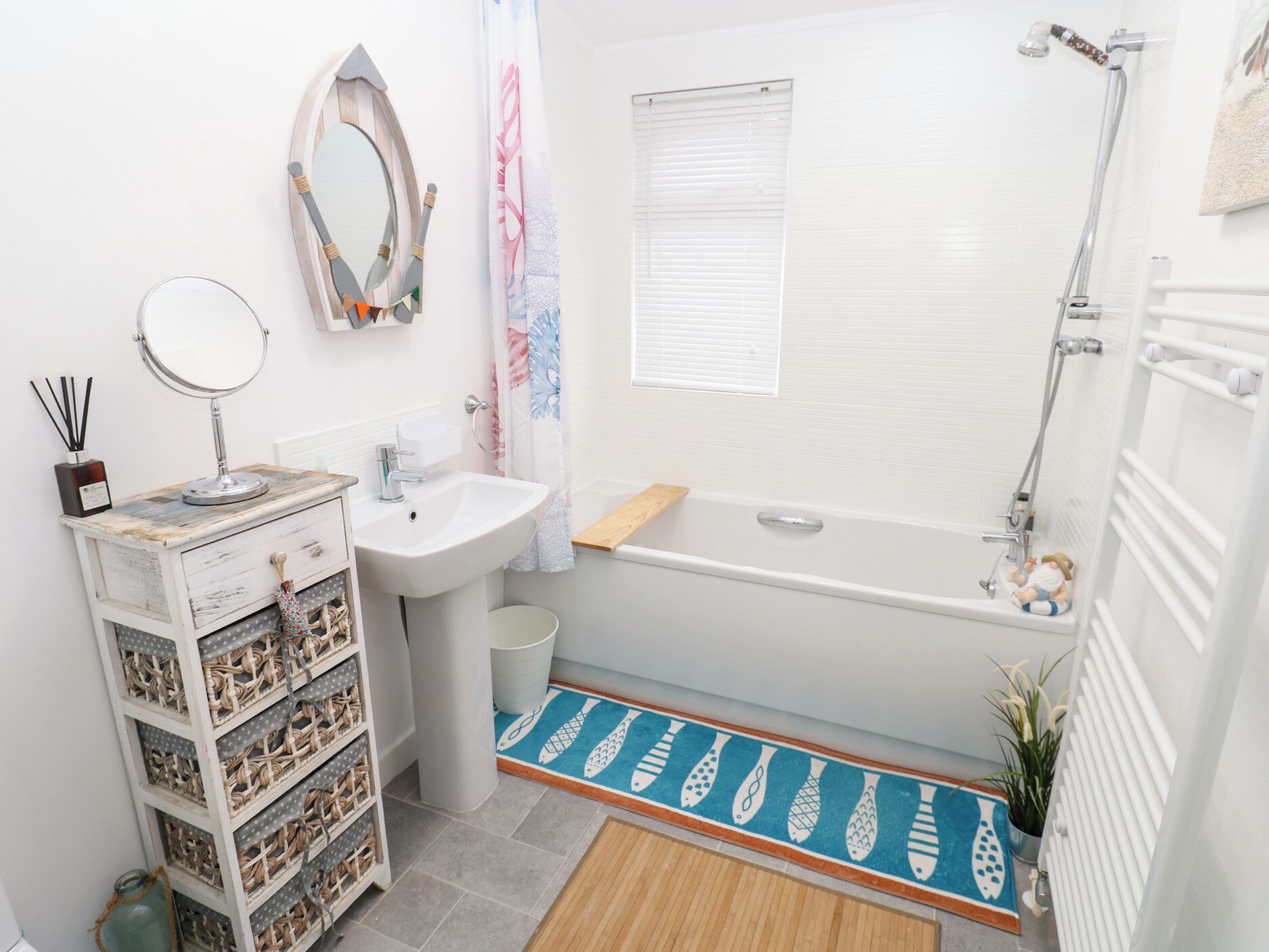 Une salle de bain avec un lavabo blanc, un miroir et une douche. On y trouve une étagère en bois et une petite plante.