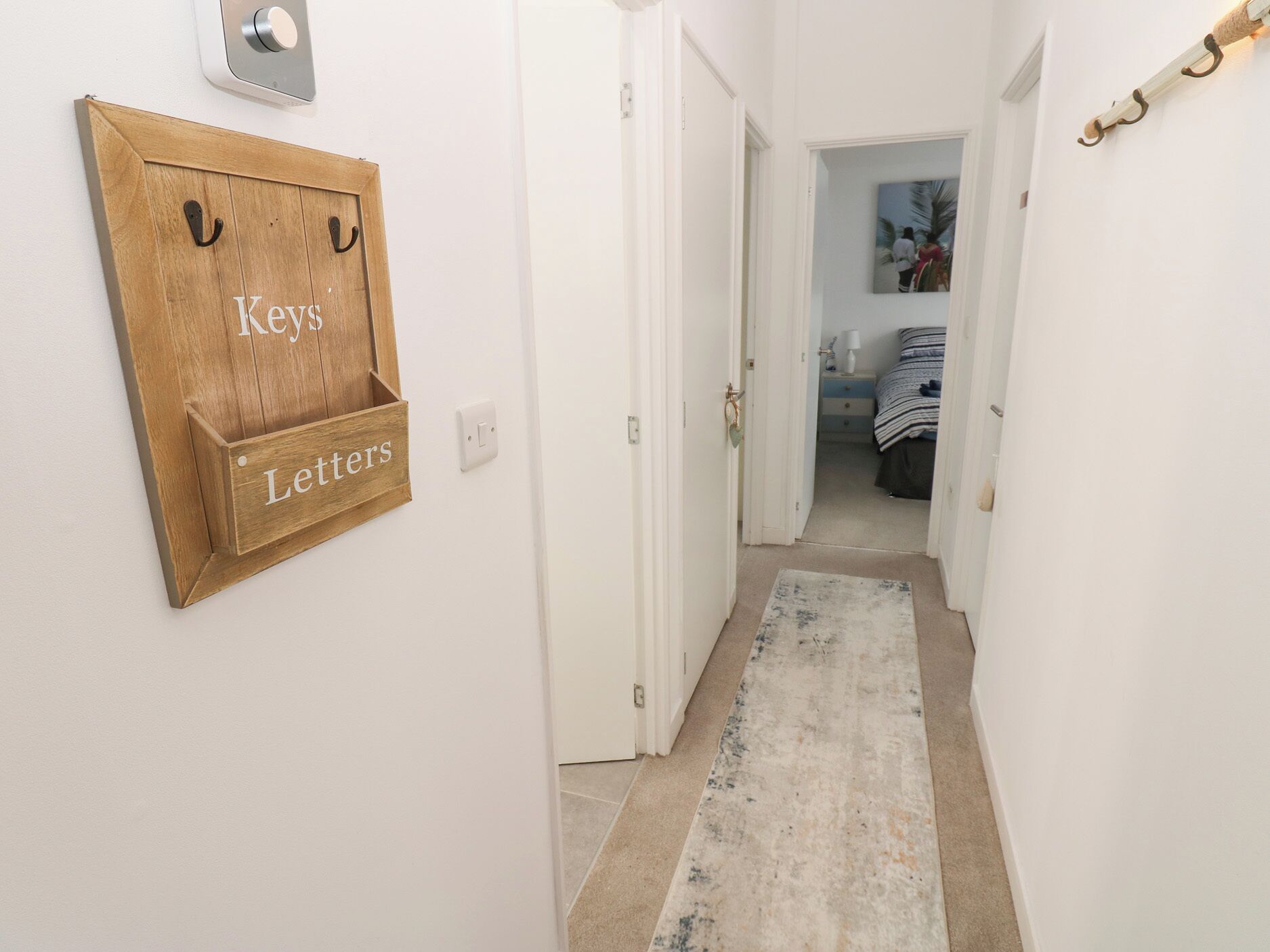 A hallway with a wooden key and letter holder on the wall, a white door, and a carpeted floor leading to a bedroom with a bed and a nightstand.