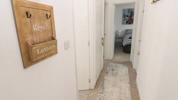A hallway with a wooden key and letter holder on the wall, a white door, and a carpeted floor leading to a bedroom with a bed and a nightstand.