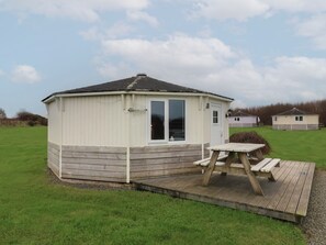 A small, single-story wooden structure with a white exterior and a large sliding glass door. It has a small deck with a picnic table and is surrounded by a grassy area. - Sandy Mouth (Bideford)