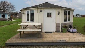 Une petite construction à un seul étage, avec une porte et des fenêtres blanches, une table de pique-nique et un petit espace jardin agrémenté d’une plante en pot.