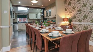 A dining area with a wooden table set for four, surrounded by brown chairs. The room has a kitchen in the background with white cabinets and a window above the sink. - The Old Post Office (York)