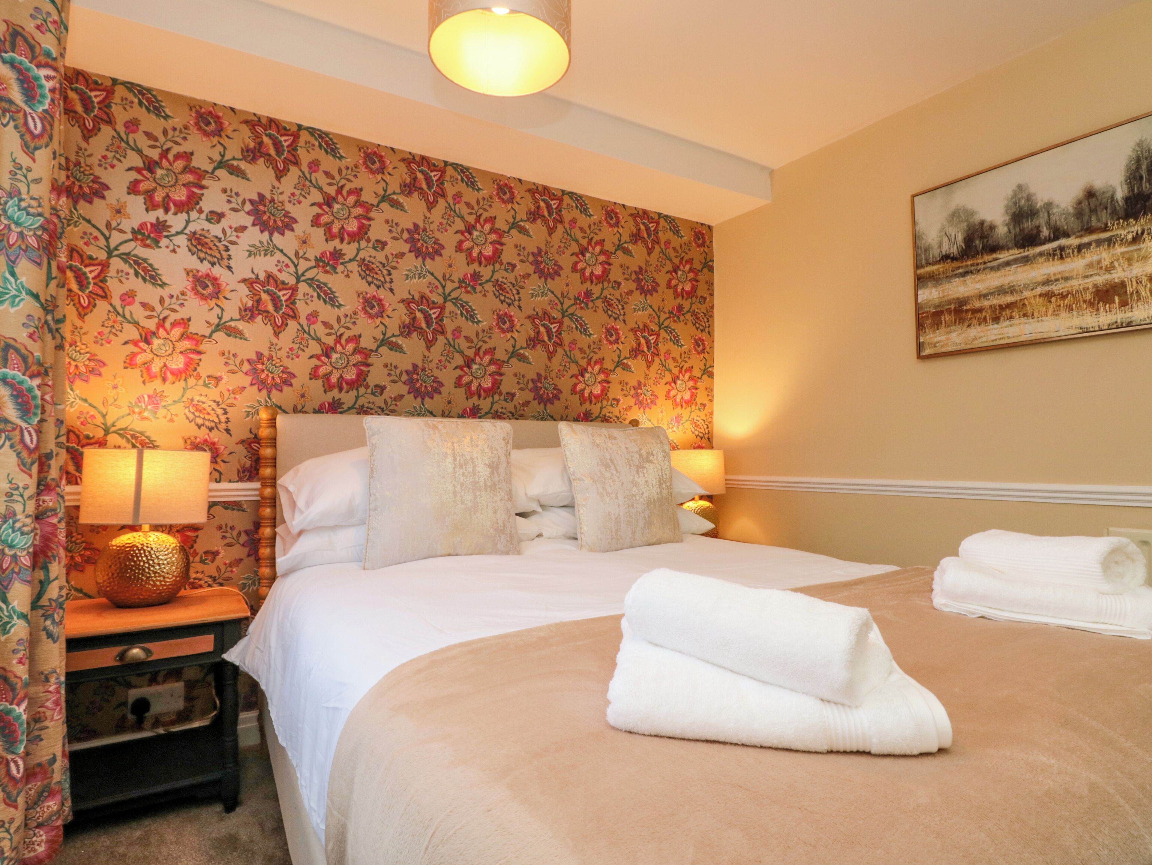 A neatly made bed with white linens and a beige duvet, a bedside lamp, and a floral-patterned wall.