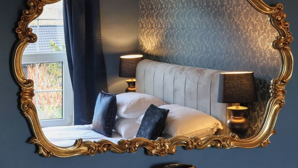 A bedroom with a large ornate mirror, two bedside lamps, a bed with pillows, and a patterned wallpaper. - The Old Post Office (York)