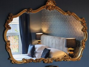 Une chambre à coucher avec un grand miroir décoratif, deux lampes de chevet, un lit avec des oreillers et un papier peint à motifs.
