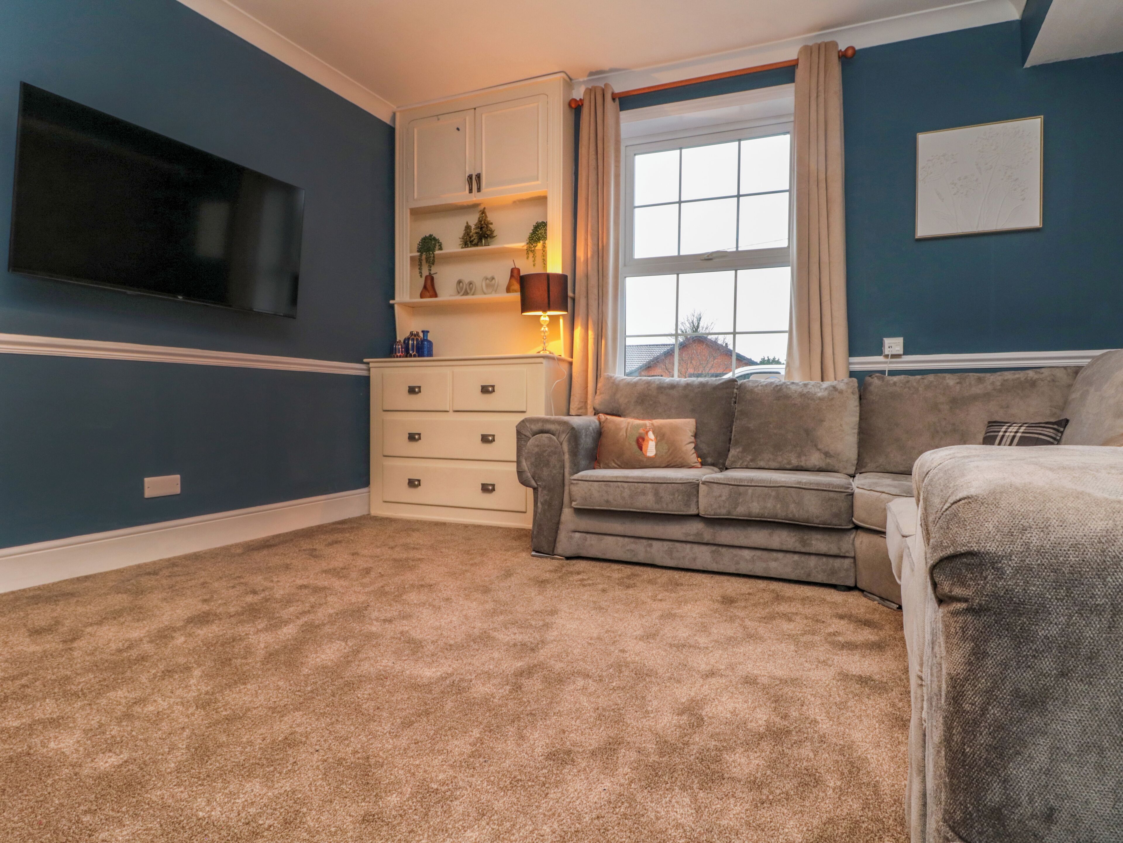 A living room with a grey sofa, a white cabinet, a flat-screen TV mounted on the wall, and a window with curtains.