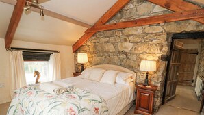 Una camera da letto con parete in pietra, soffitto in legno, un letto con biancheria da letto a fiori, due lampade da comodino e una finestra con tende.