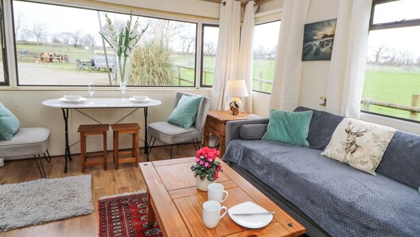 A cozy living room with a sofa, a coffee table, and a dining table. There are chairs around the dining table, and a window with a view of the outdoors. - Upper Calffield Caravan (Haverfordwest)