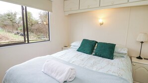 A neatly made bed with a white and green floral bedspread, a white towel, and a nightstand with a lamp. - Upper Calffield Caravan (Haverfordwest)
