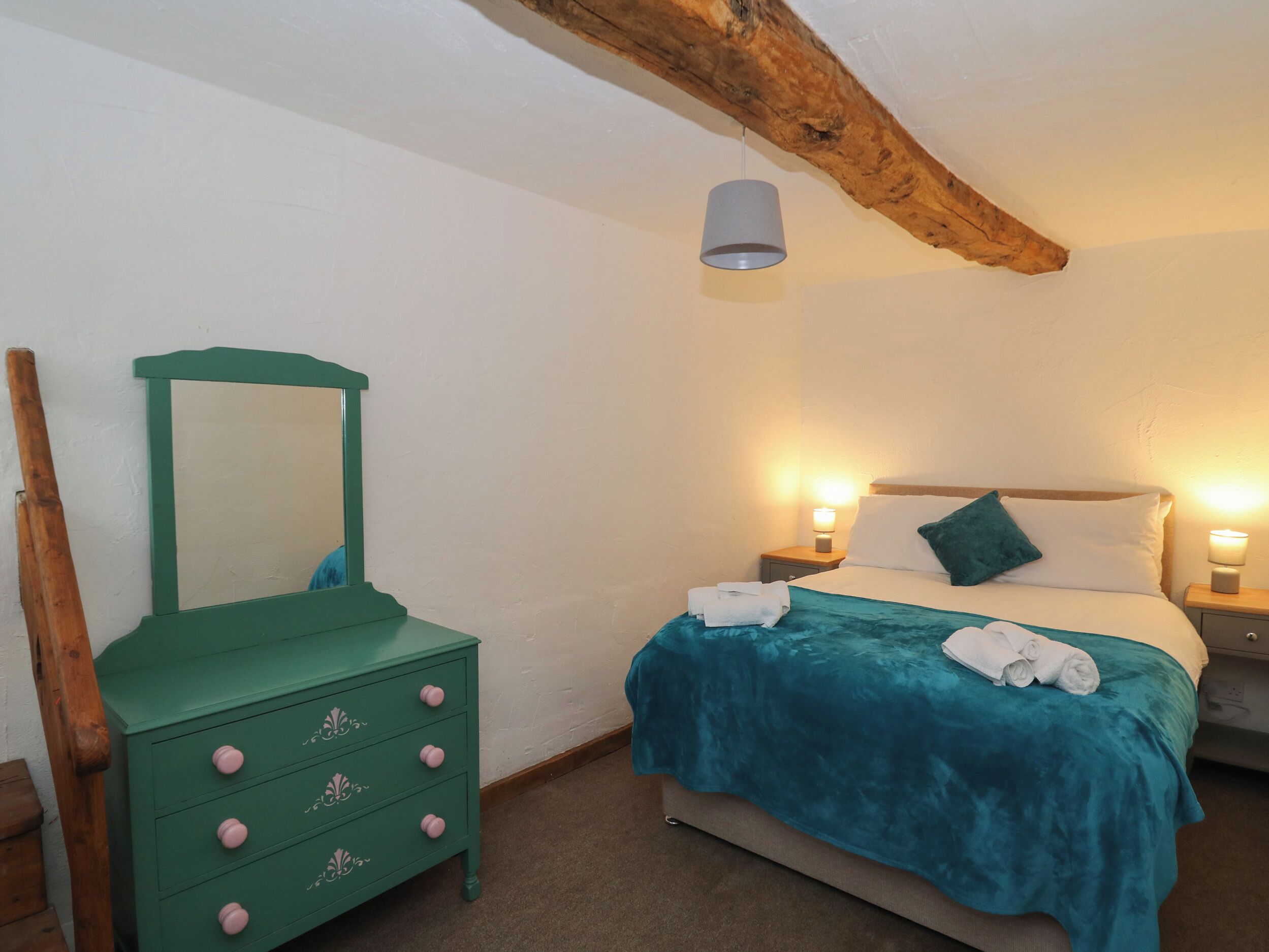 A bedroom with a bed, a green dresser with pink knobs, a wooden beam ceiling, and a hanging light fixture.
