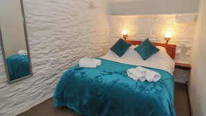 A bedroom with a stone wall, a bed with teal bedding, two pillows, a nightstand with a lamp, and a mirror on the wall. - Y Beudy (Llanybydder)