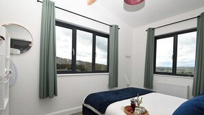 Una camera da letto con due ampie finestre, un letto con coperta blu, un comodino con cestino di frutta e una tazza, e un'armadio bianca.