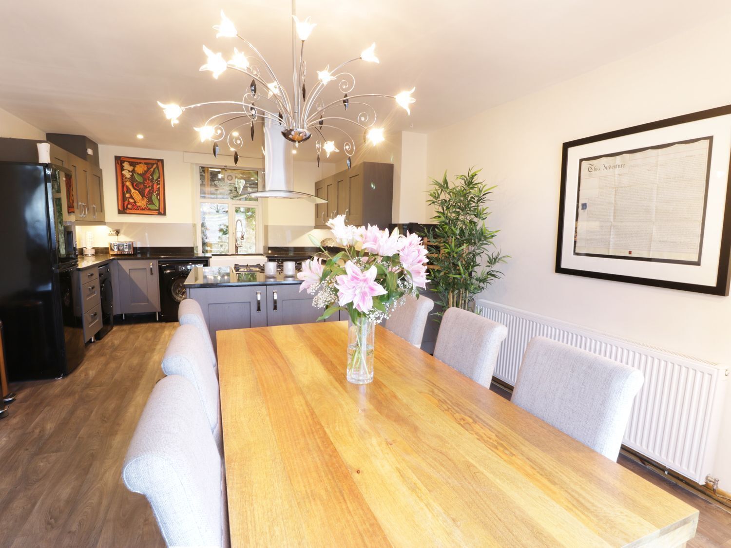 A modern kitchen with a wooden dining table, a chandelier, and a framed menu on the wall.
