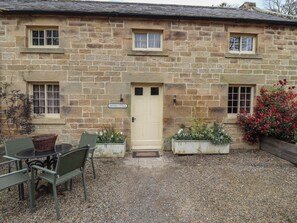 A stone cottage with a sign reading "Grooms Cottage," a front door, and a gravel patio with a table and chairs.