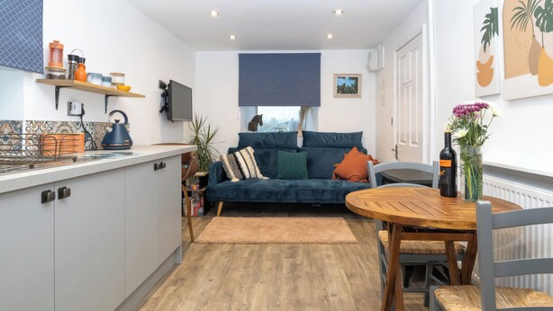 A modern kitchen with a built-in stove, a dining area with a wooden table and chairs, a blue sofa with cushions, and a large window with a view. - Duck Tavern Annexe (Rhosgoch)