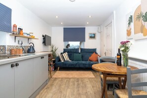 A modern kitchen with a built-in stove, a dining area with a wooden table and chairs, a blue sofa with cushions, and a large window with a view. - Duck Tavern Annexe (Rhosgoch)