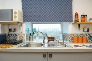 A kitchen with a microwave, sink, and window with a view. There are utensils and containers on the counter. - Duck Tavern Annexe (Rhosgoch)