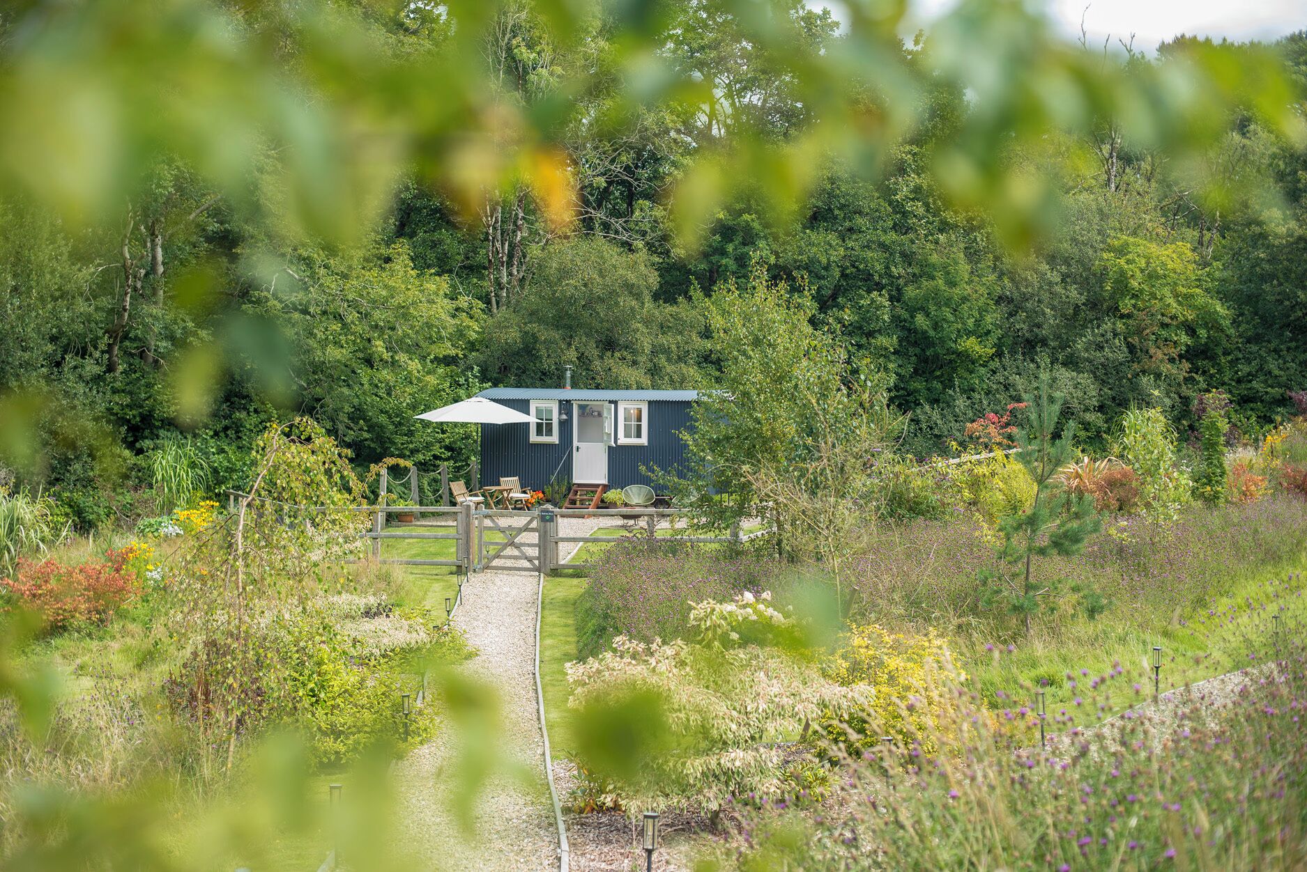 A small, single-story cabin with a metal roof, surrounded by a garden and a gravel path leading to the entrance.