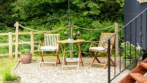 Area salotto esterna in legno con due sedie e un tavolo, delimitata da una recinzione in corda e affacciata su una vegetazione rigogliosa.