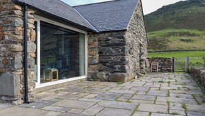 A stone building with a large glass window, a stone pathway, and a view of a green hillside. - Beudy Cefn (Porthmadog)