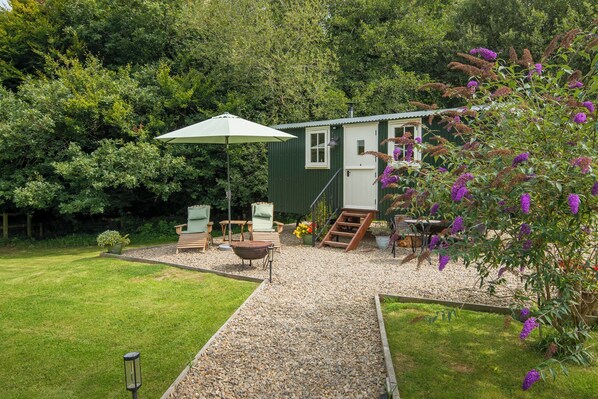 A small, green house with a gravel patio, two wooden chairs, a table, and an umbrella. - Collen (Llanarth)