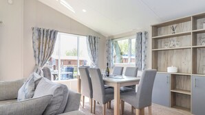 Een moderne woonkamer met een bank, eettafel en stoelen. Er is een groot raam met gordijnen, een boekenkast en uitzicht op buiten.