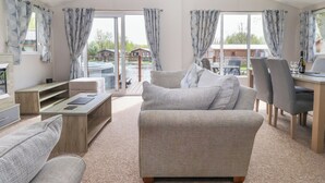 Een ruime woonkamer met een grote bank, een houten salontafel en een eethoek met stoelen en een tafel. Er zijn grote ramen met gordijnen, en uitzicht op een jacuzzi en gebouwen.