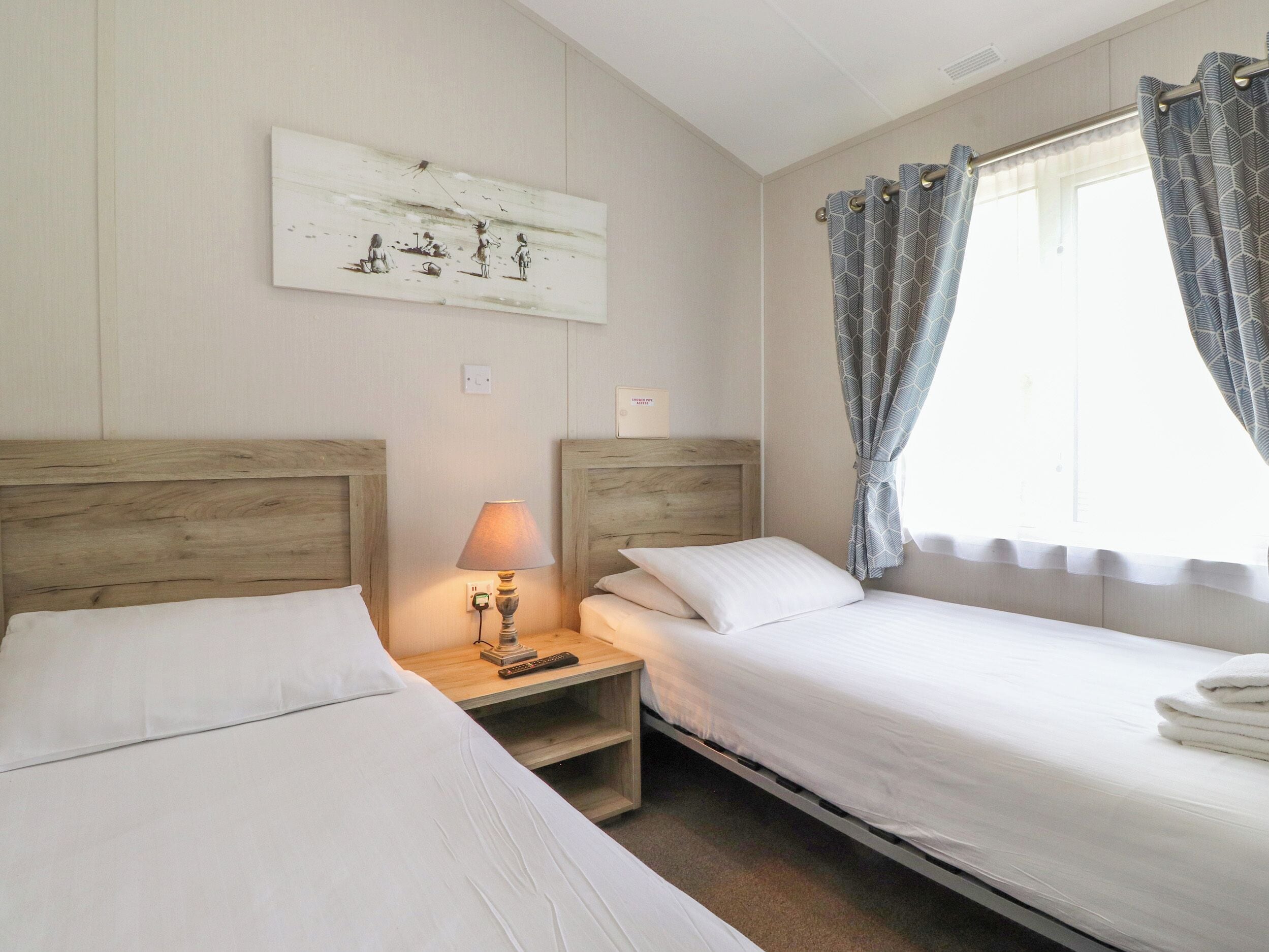 A hotel room with two single beds, a nightstand with a lamp, a painting on the wall, and a window with patterned curtains.