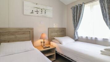 A hotel room with two single beds, a nightstand with a lamp, a painting on the wall, and a window with patterned curtains.