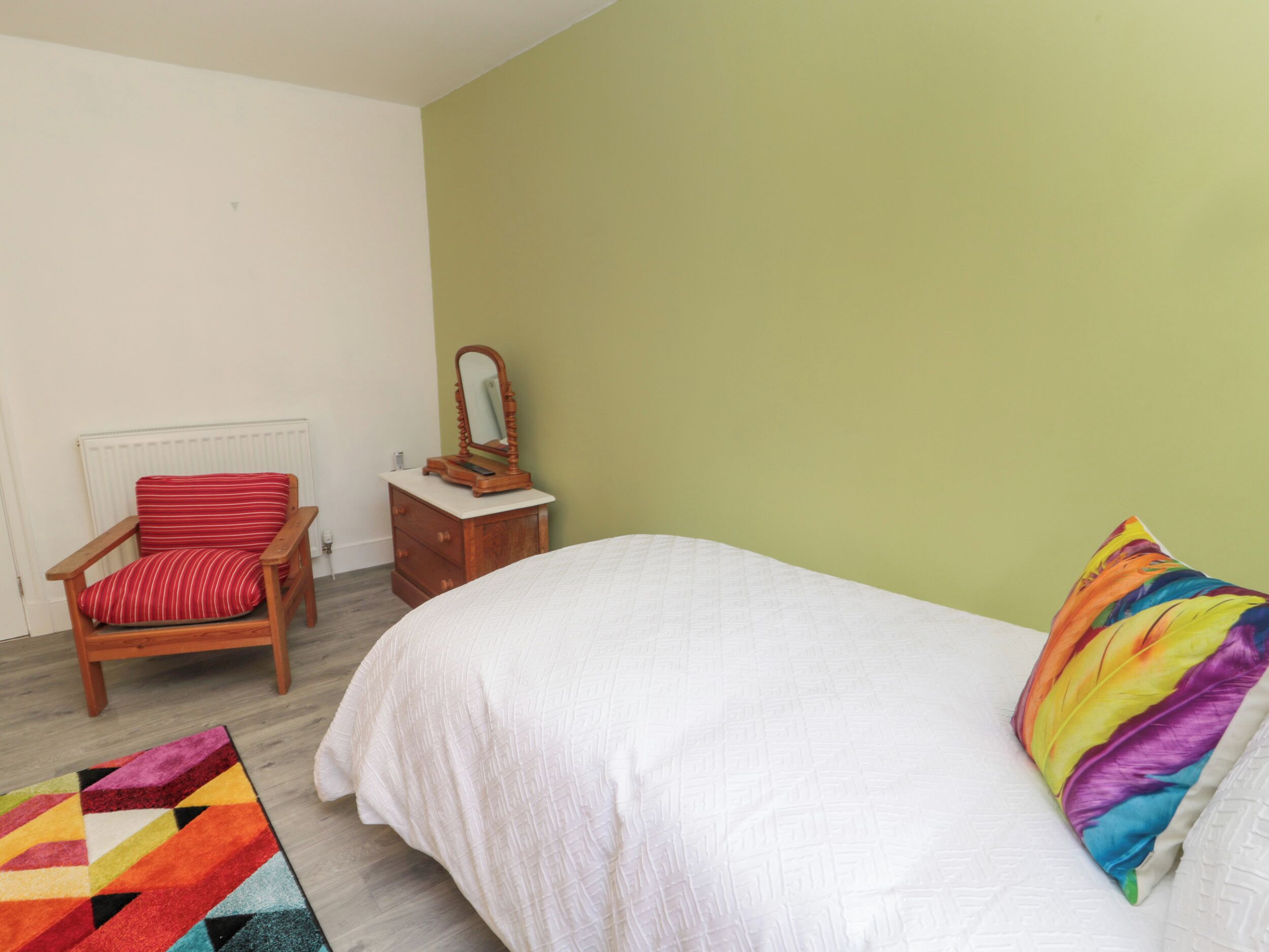Ein Schlafzimmer mit einem Bett, einem Sessel mit gestreifter Sitzkissen, einer Holzkombination mit Spiegel und einem farbenfrohen Teppich.