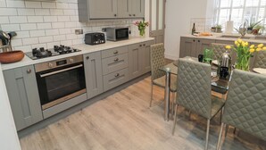 A modern kitchen with grey cabinets, a stainless steel range hood, a built-in oven, and a dining area with a glass table and cushioned chairs. - The Old Factory House (Berwick-upon-Tweed)