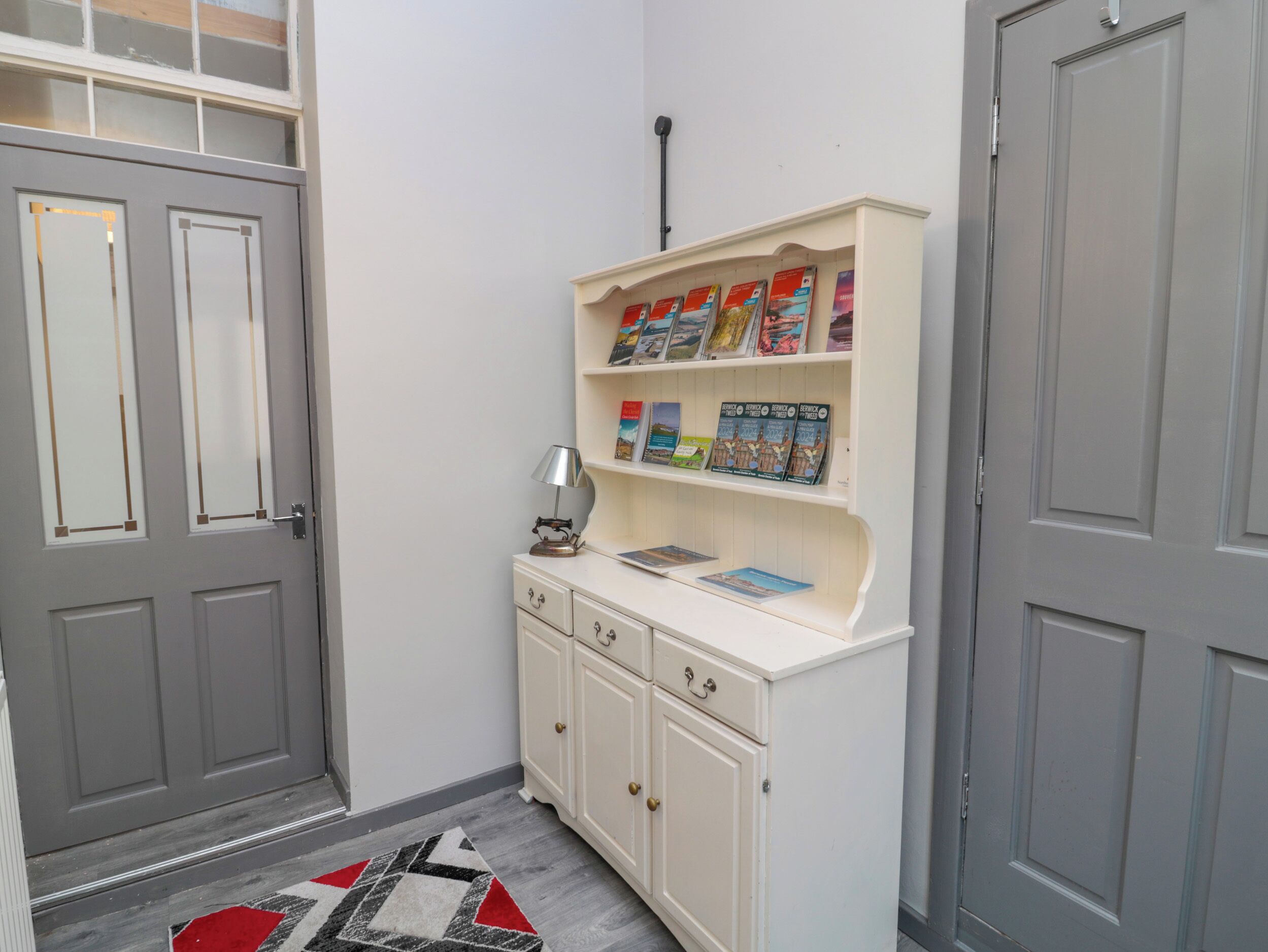 Un pasillo con puertas grises, un armario blanco lleno de libros y una alfombra estampada.