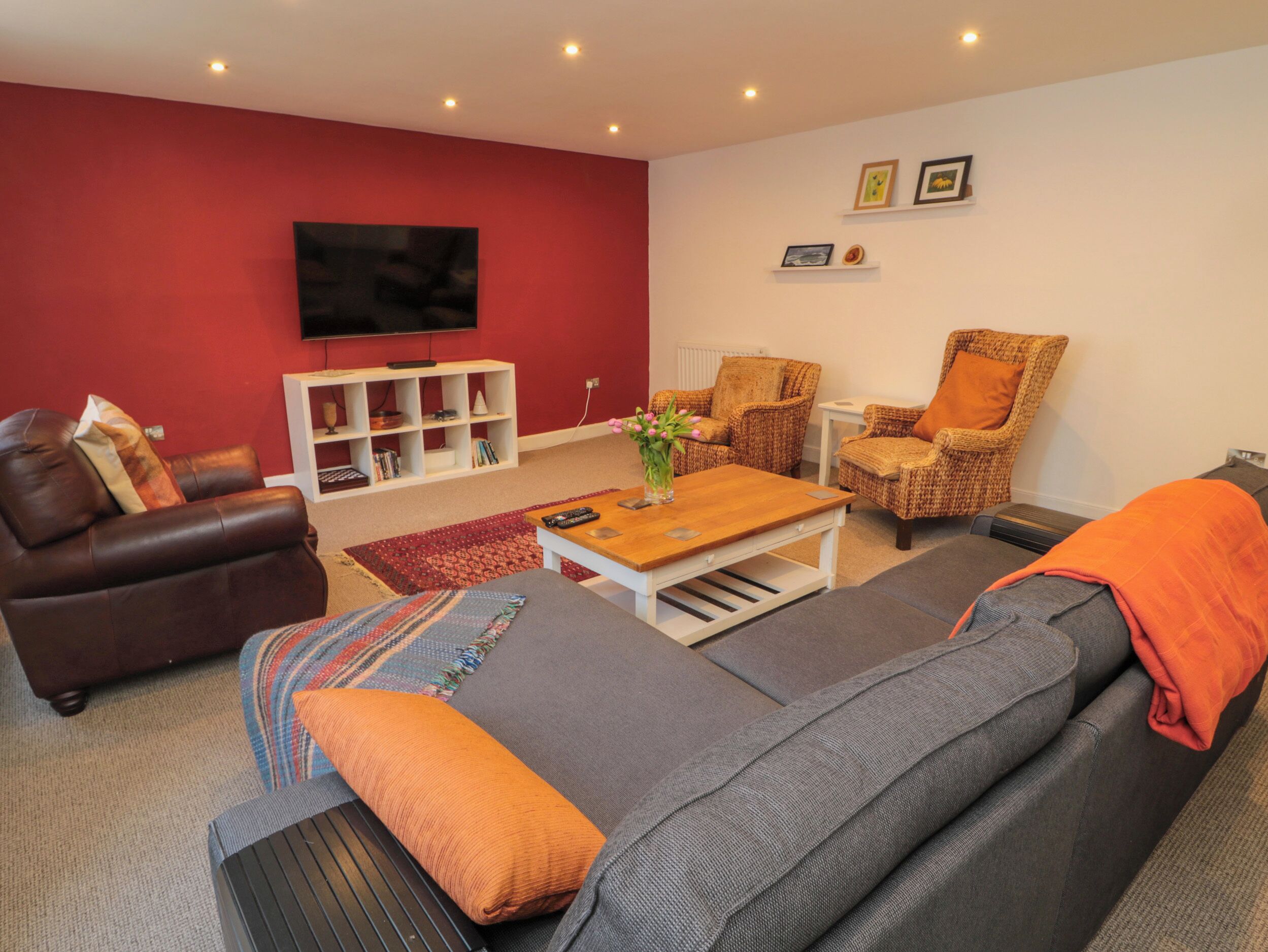 A living room with a brown leather sofa, grey sofa, wicker chairs, a wooden coffee table, and a flat-screen TV mounted on the red wall.