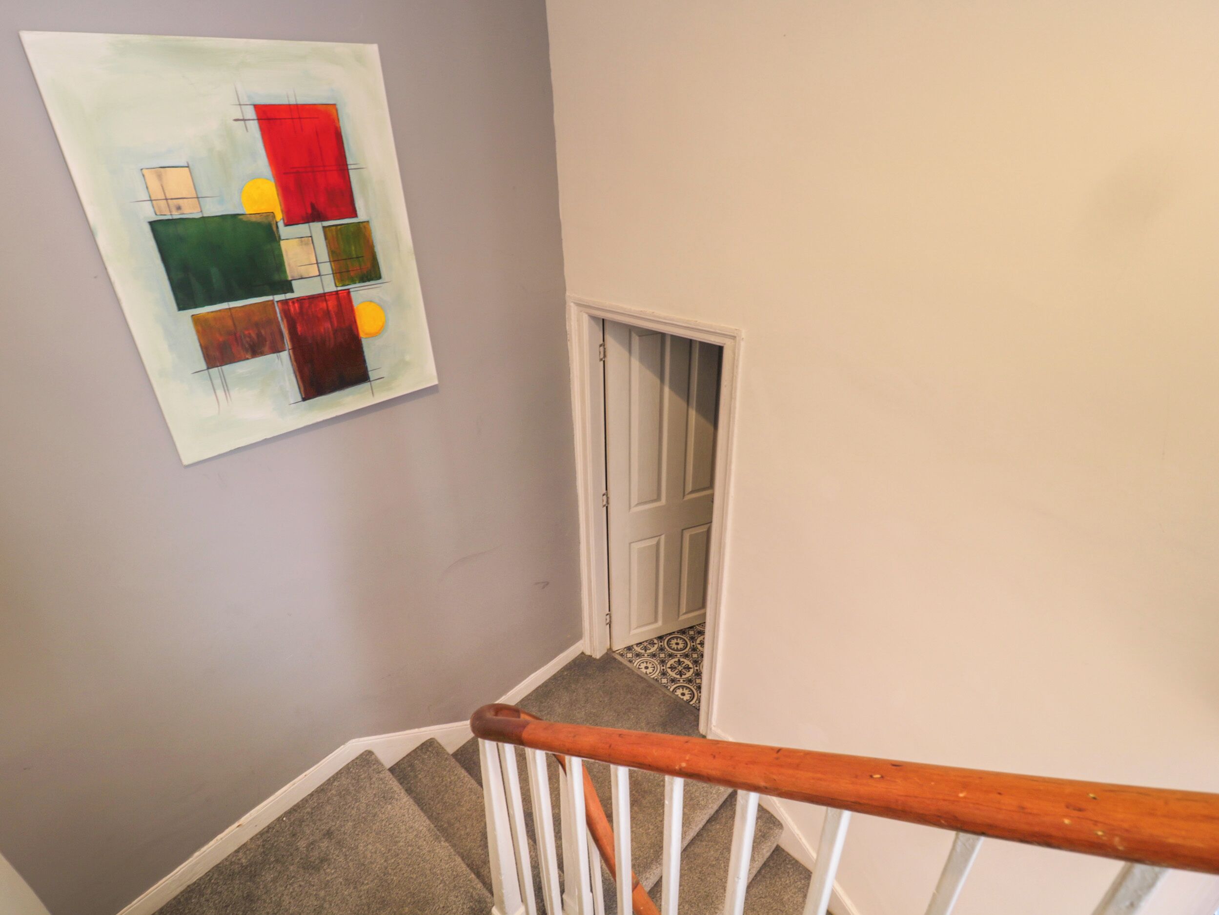 Een trap met een houten leuning, een deur die naar een kamer leidt en een kleurrijk abstract schilderij aan de muur.