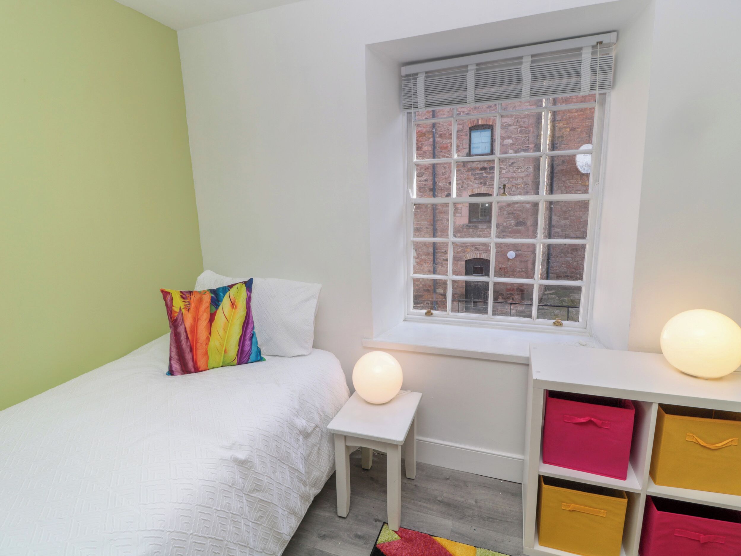 Ein Schlafzimmer mit einem Bett, einem kleinen Tisch, einem Fenster mit Blick auf ein Backsteingebäude und Aufbewahrungsboxen.