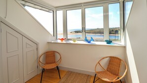 Ein Zimmer mit schrägem Dach, großen Fenstern mit Blick auf einen Strand, zwei Korbstühle und ein kleines Regal mit Dekoration.