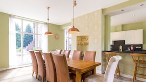 A dining area with a wooden table and chairs, a kitchen area with cabinets and a countertop, and a large window with curtains. - The Coach House (Belford)