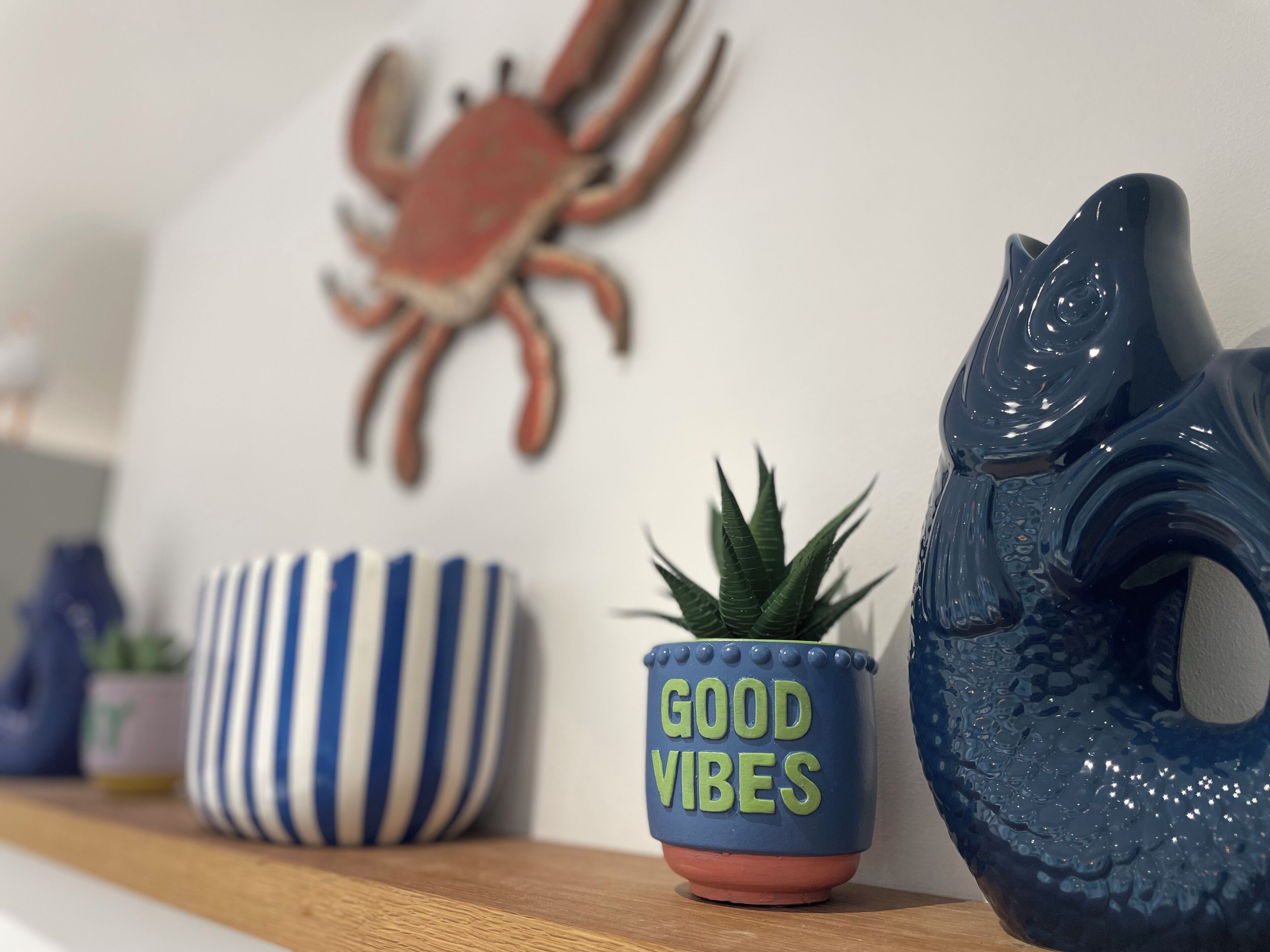 A shelf with decorative items including a blue ceramic crab, a striped bowl, a small potted plant, and a ceramic cup with "GOOD VIBES" text.