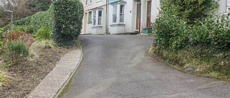 Une rue résidentielle étroite, bordée de maisons des deux côtés, avec un trottoir en béton et divers arbustes et plantes.