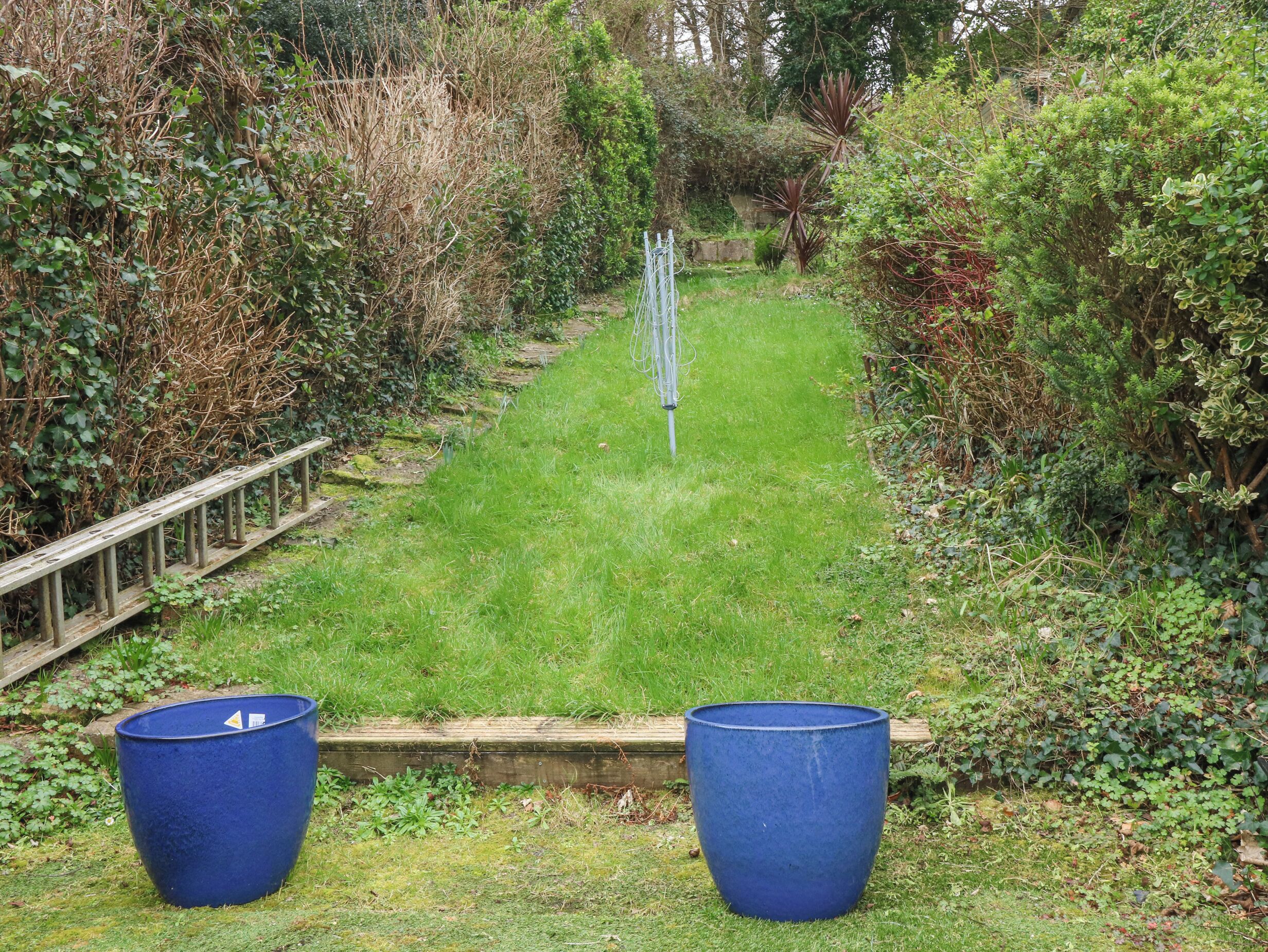 Un jardin avec une clôture en bois, un sentier recouvert d’herbe et deux grands bacs à fleurs bleus.