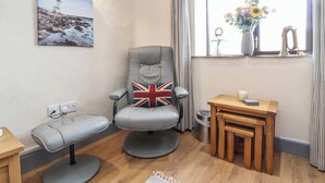 A corner room with a grey armchair, a bench, a small wooden table, a window with curtains, a wall-mounted picture, and a trash bin. - Badgers Sett (Bude)