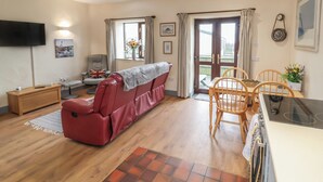 A living room with a red sofa, a wooden dining table with chairs, a TV mounted on the wall, and a sliding glass door leading to an outdoor area. - Badgers Sett (Bude)