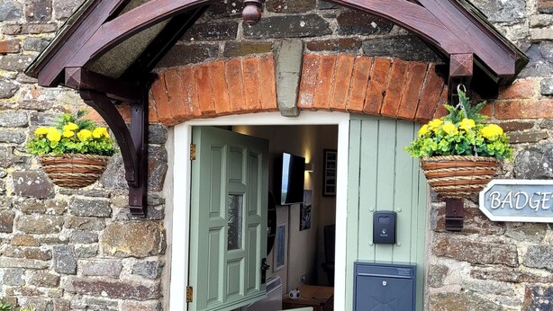 A rustic building entrance with a green door, a sign reading "BADGE", and two baskets of yellow flowers. - Badgers Sett (Bude)