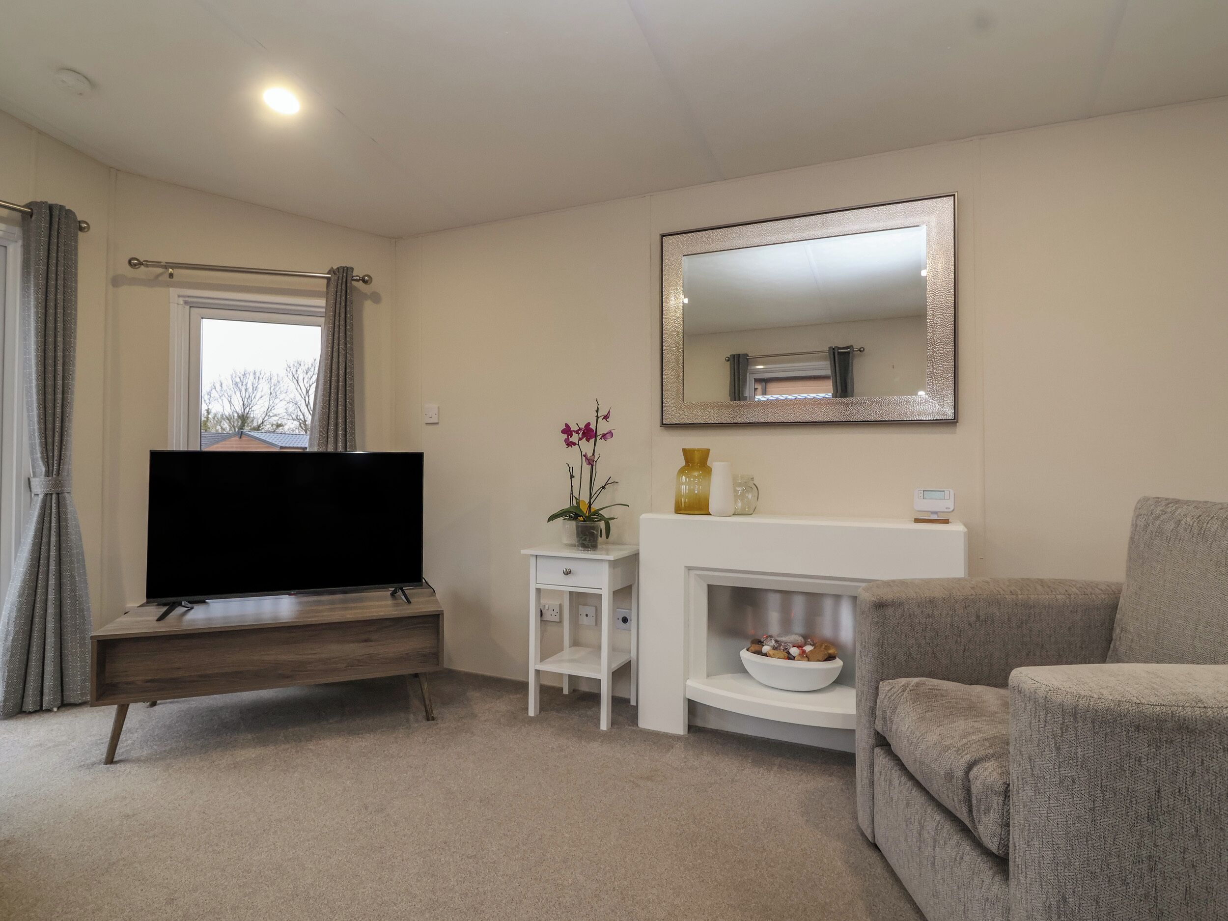 A living room with a flat-screen TV on a wooden stand, a white fireplace with a mirror above it, a grey sofa, and a small white side table with a vase of flowers.
