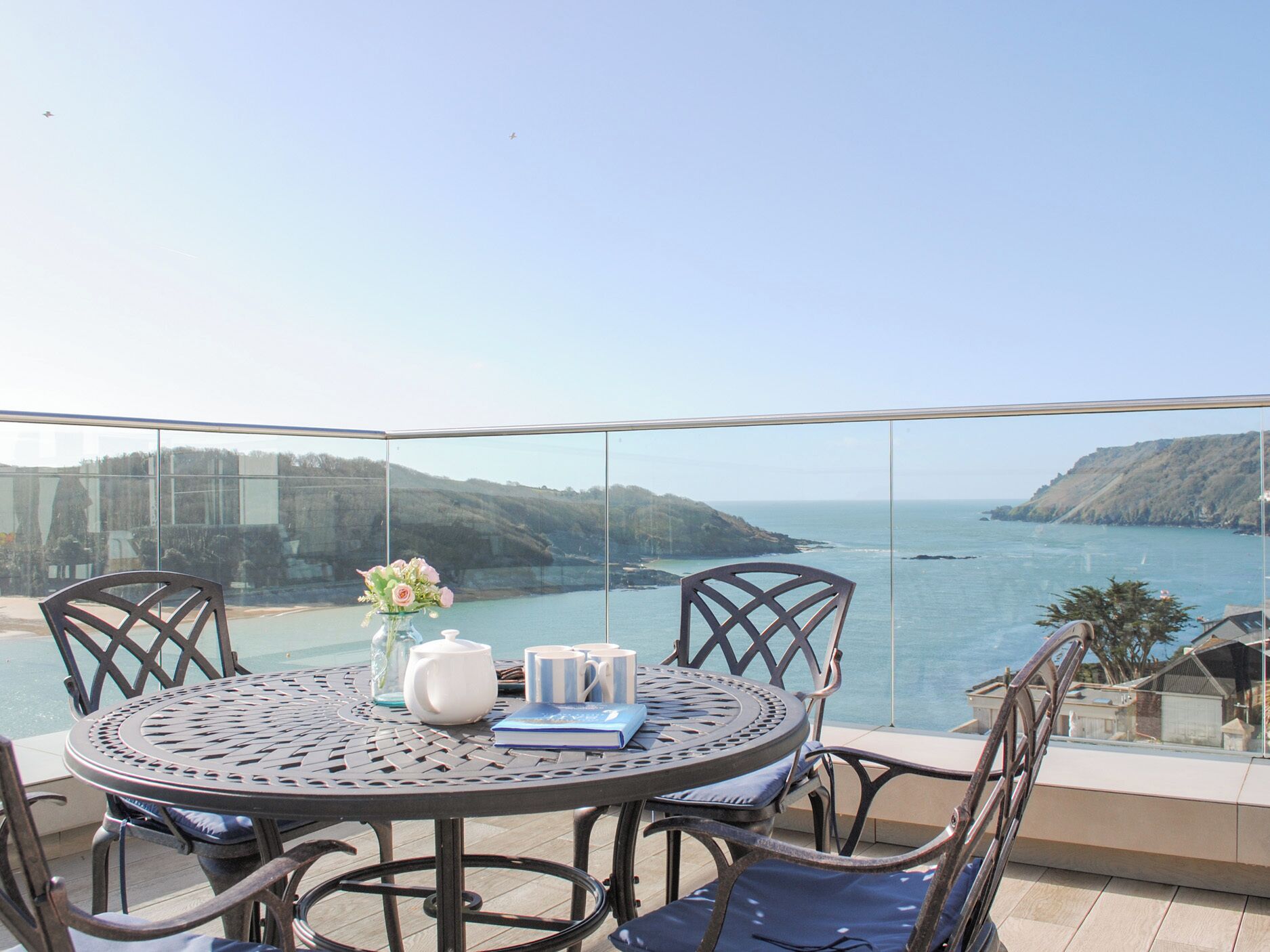 A round metal table with chairs on a balcony overlooking a coastal view.