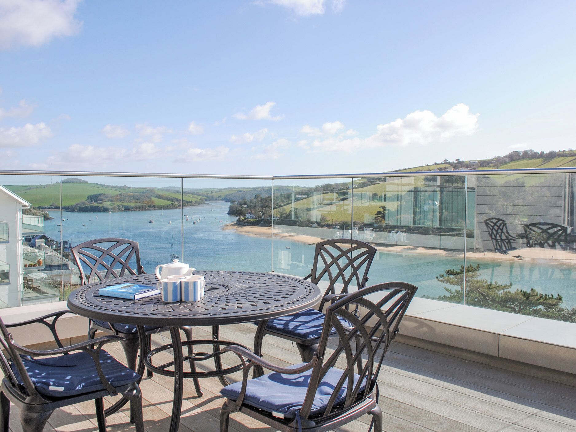 A round table with chairs and a glass balustrade overlooking a body of water with boats, a beach, and hills in the distance.